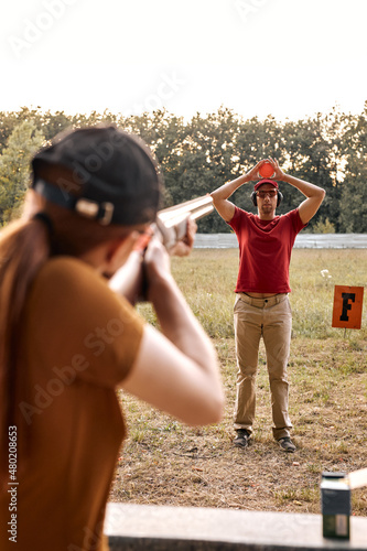 Rear view on woman in cap and goggles aiming rifle at target pigeon clay in hands of male instructor, ready to shoot,concentrated, practice and training in outdoor range. focus on man in background