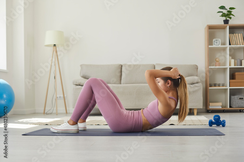 Woman exercising on rubber mat on floor in living room during routine fitness workout at home. Side view of fit young Caucasian girl in sports clothes training her abs by doing sit ups or crunches