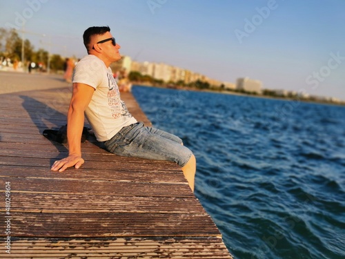  Portrait of a young, athletic man sitting on the edge of the embankment by the sea. He enjoys the sunset and the surrounding nature. Travel, vacation, Greece