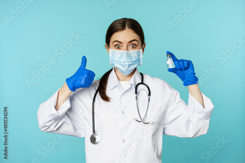 Portrait of smiling doctor, medical personel in face medical mask and rubber gloves, showing thumbs up and coronavirus, omicron vaccine, standing over blue background