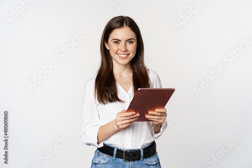 Portrait of smiling corporate woman looking at digital tablet, working, standing over white background