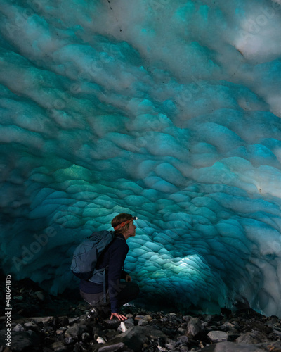 Adventurous Ice Climber Woman walking in an ice cave on a glacier in Alaska. Glacier is receding and melting due to global warming. Dark blue ice and headlamp