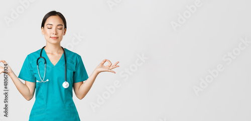 Covid-19, healthcare workers and preventing virus concept. Calm and patient asian female doctor, nurse in scrubs staying relaxed, meditating with eyes closed and happy smile, white background