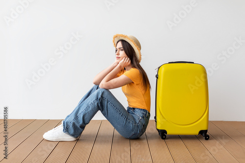 Upset female tourist sitting near suitcase and looking away at free space over light wall background