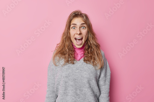 Happy positive woman amazed by big sales reacts on something shocking keeps mouth widely opened dressed in casual grey jumper isolated over pink background screams loudly. Human reactions concept