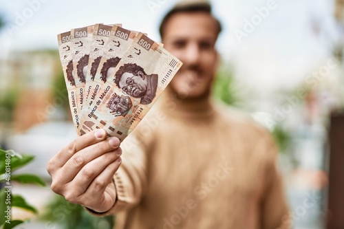 Young caucasian man smiling happy holding mexican pesos banknotes at the city.