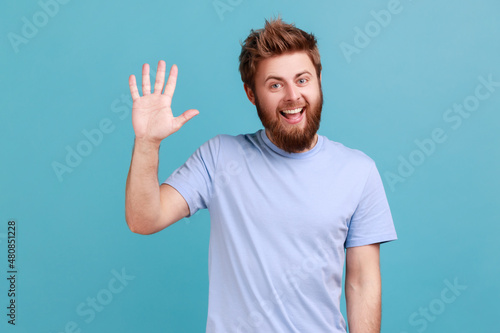 Portrait of positive bearded man greeting you rising hand and waving, saying hi, glad to see you, looking at camera with toothy smile. Indoor studio shot isolated on blue background.