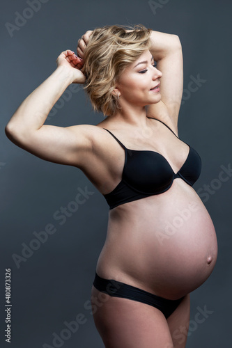 Pregnant woman in black underwear. Beautiful young smiling blonde woman keeps her hands behind her head. Happiness in anticipation of a baby. Gray background. Vertical.