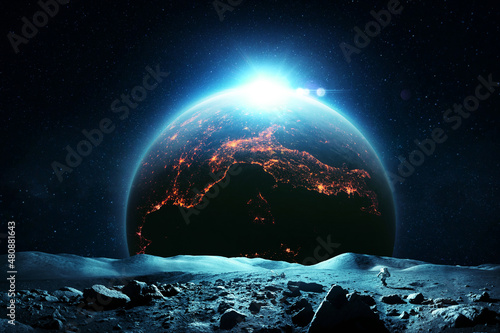Astronaut man in a space suit and a lunar rover exploring the surface of the moon. Beautiful moon and planet Earth with night city lights and dawn rays. Amazing space wallpaper