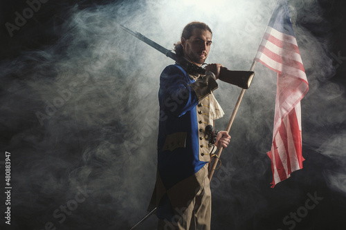 American revolution war soldier with flag of colonies and musket gun over dramatic smoke background. 4 July independence day of USA concept photo composition: soldier and flag.