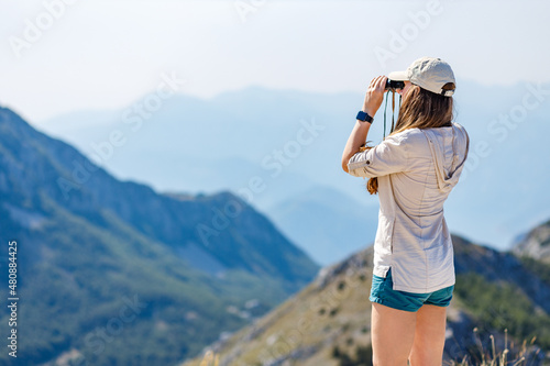 Young woman watching mountain landscape through binoculars at the view spot on tourist trail.