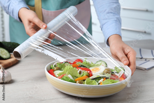 Woman putting plastic food wrap over bowl of fresh salad at wooden table indoors, closeup