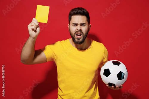 Irritated young bearded man football trainer in t-shirt hold soccer ball hold soccer ball show yellow card isolated plain dark red background studio portrait. People sport leisure lifestyle concept