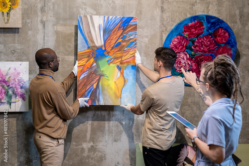 Two young men hanging abstract painting on wall while preparing for presentation of new artwork collection