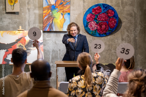 Mature male auctioneer in formalwear standing by tribune and pointing at one of people with auction paddles