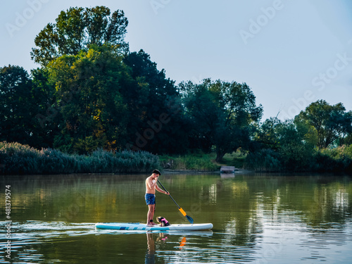 Young person stand on paddle board with his small yorkshire terrier and swimming on a river in a sunny day. Stand up paddle boarding or paddling is awesome active recreation in nature.