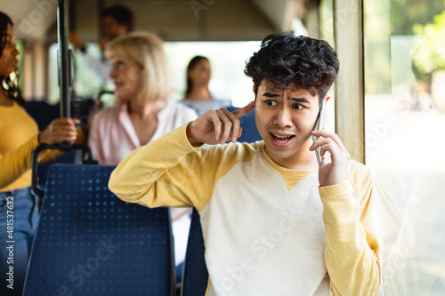 Confused asian guy talking on phone holding finger in ear