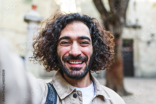 Joyful male with dark curly hair looking at camera and taking self portrait while standing on street with buildings on blurred background