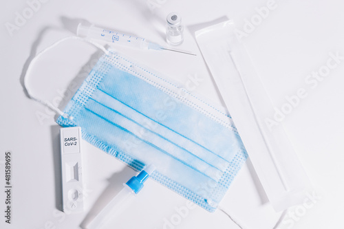 COVIC-19 coronavirus vaccine concept and elements to perform an antigen test against the virus. On the white table is a complete kit of blue surgical mask, diluent tube, swab and vaccine vial...