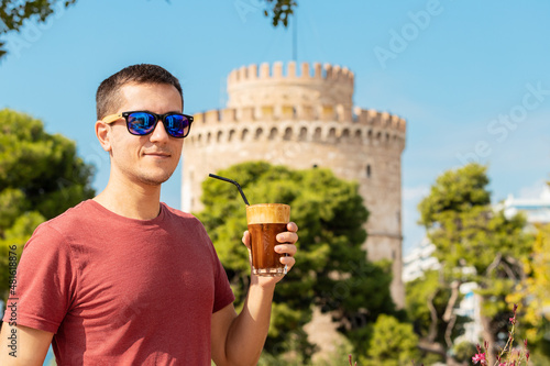 A happy smiling young man drinks traditional Frappe coffee against the background of the famous White Tower in the city of Thessaloniki in Greece.
