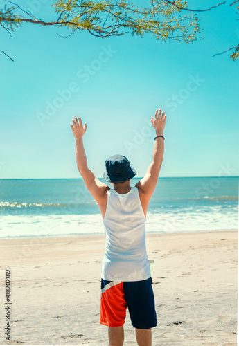 man with hat from back on the beach, young man from back raising his hands on the beach.