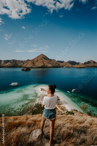 A young female traveler is exploring tropical island in Komodo, Indonesia. She is wearing a casual outfit and overlooks lagoon bag, boat and hills in the distance