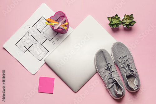 Laptop with drawings, office supplies and sports shoes on color background