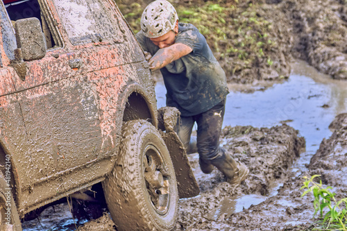 A man wearing a helmet pushes an orange 4x4 off-road car through the mud. Extreme off-road competition on an off-road vehicle. The concept of adventure travel.