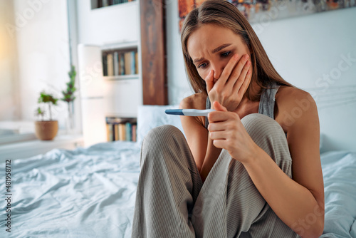 Excited young girl in home clothes sitting on the bed and looking sadly at the pregnancy test covering her mouth with her hand in surprise.