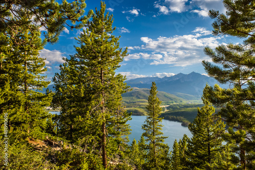 Colorado lake with green pines in the foreground, mountains in the background and white clouds on blue sky.