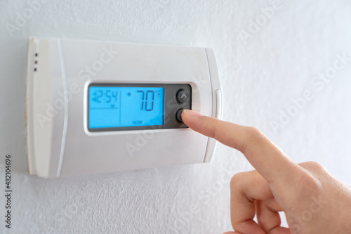 A woman is pressing the down button of a wall attached house thermostat with digital display showing temperature 70 degree Fahrenheit for heating, cooling, electricity and gas saving