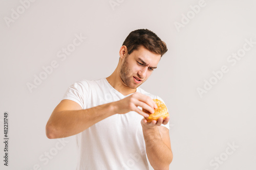 Portrait of dissatisfied young man with disgust picking bud of bad burger on white isolated background. Studio shot of handsome bearded male holding in hands unhealthy delicious hamburger.