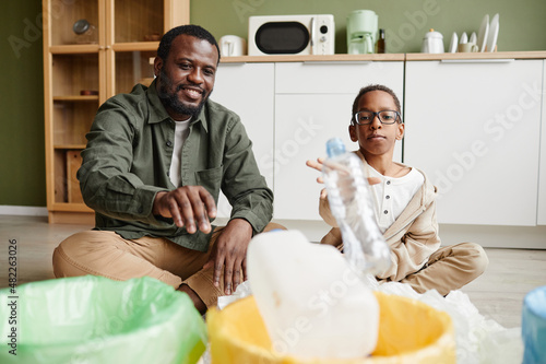 Full length portrait of happy African-American father and son putting plastic in recycling bins at home, copy space