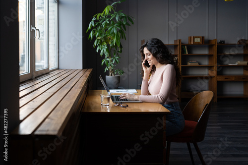 Happy smiling female entrepreneur having pleasant mobile conversation with client or customer, young successful business woman working with online order on laptop computer and talking via cellphone