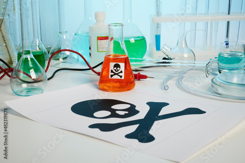 Death printed symbol on a chemical table. Poison fluids in toxicology laboratory. Photo contains danger paper sheet, test tubes, orange colored liquid in chemical flask, some retorts, electrodes