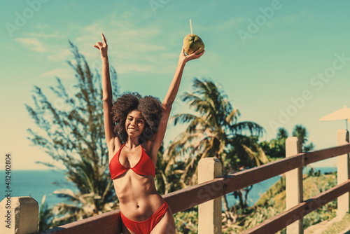 A dazzling happy young Brazilian female with curly fluffy afro hair is enjoying a sunny day on a tropical beach of a resort, standing in a red bikini next to a fencing with hands raised and a coconut