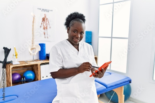 Young african american woman wearing physiotherapist uniform using touchpad at clinic
