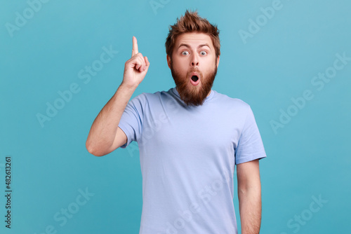 Eureka. Portrait of clever smiling bearded man pointing finger up, has good idea genius plan, inspiration and creativity, keeps mouth open. Indoor studio shot isolated on blue background.