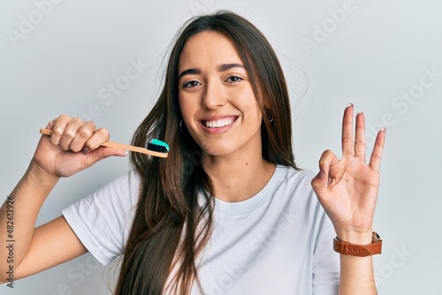Young hispanic girl holding toothbrush with toothpaste doing ok sign with fingers, smiling friendly gesturing excellent symbol