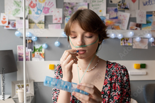 Teenage girl with oxygen nose pipe taking pills and looking at medicaments in long plastic container in hospital ward