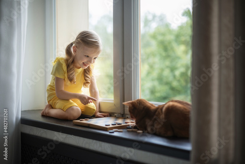 Beautiful girl on the windowsill plays checkers with a cat