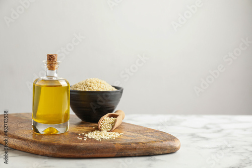 Wooden board with bottle of sesame oil and seeds on white marble table. Space for text