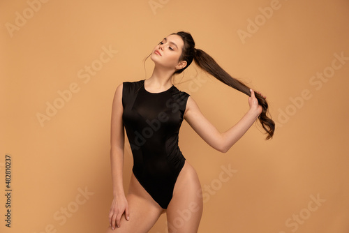 Young beautiful woman dressed seductive black bodysuit standing studio isolated on color background holding hair ponytail looking away