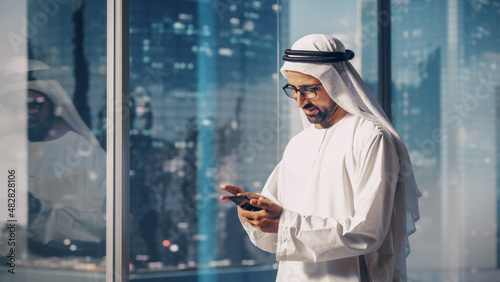 Successful Muslim Businessman in Traditional White Outfit Standing in His Modern Office, Using Smartphone Next to Window with Skyscrapers. Successful Saudi, Emirati, Arab Businessman Concept.