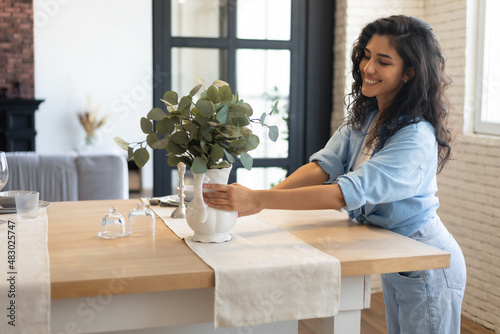 Beautiful young brunette woman arranging plants in vase, decorating dining room table, creating cozy atmosphere at home