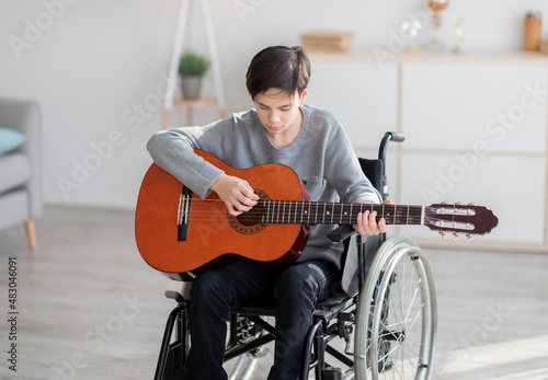 Focused disabled teen boy in wheelchair playing guitar, creating music at home. Stay home hobbies during covid lockdown