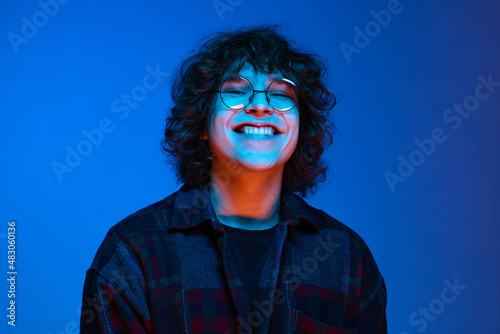 Portrait of young smiling man, student in black hoodie isolated on dark blue background in neon light. Concept of emotions, facial expression, youth, aspiration, sales