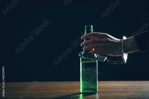 Alcohol addiction. Woman handcuffed to bottle of beer against black background, closeup. Space for text