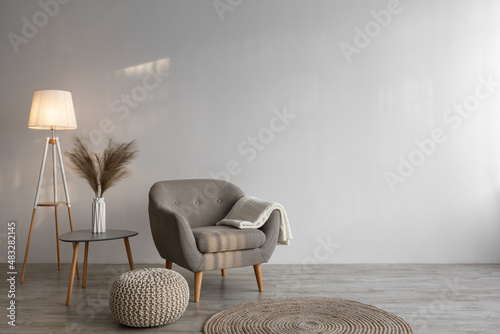 Retro armchair with plaid, luminous lamp, dry plants in vase on table, ottoman and round carpet