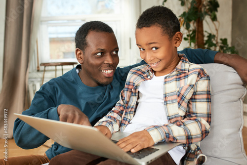 Sly-looking dark-skinned boy kid holding opened laptop on knees, to play games, browse web-sites, smiling sitting next to his eldest brother of 20s, looking at him with smile while pointing at screen